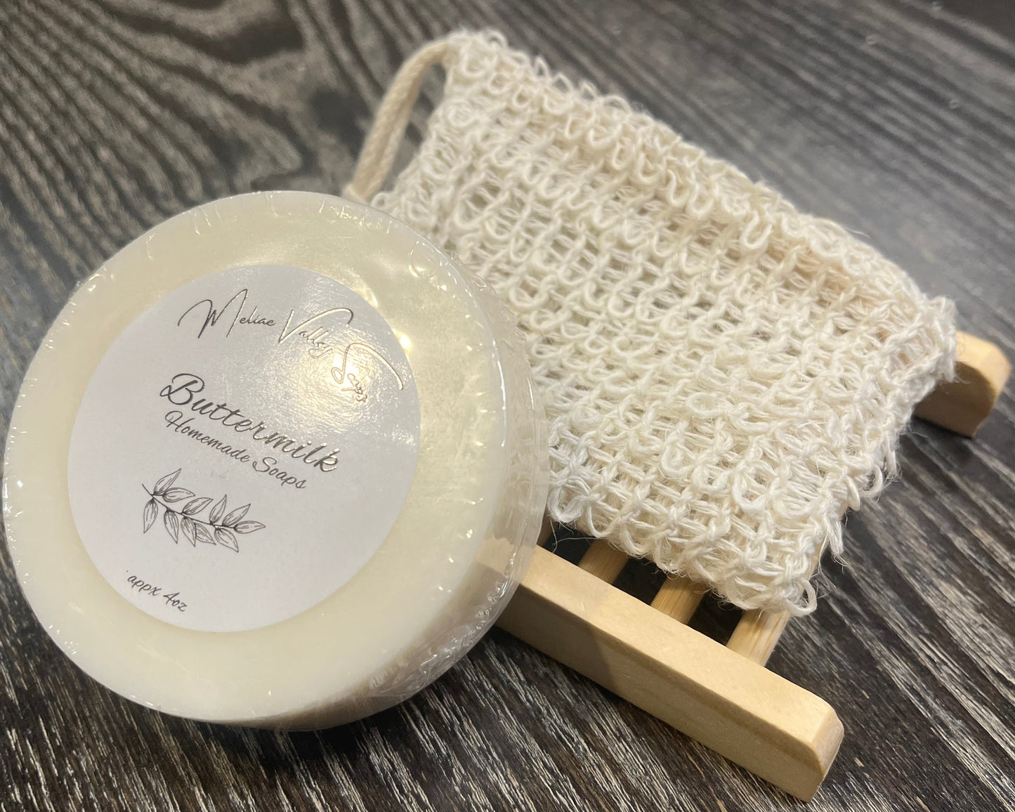 A round white soap with a wooden stand and an exfoliating bag on a dark wooden surface. 