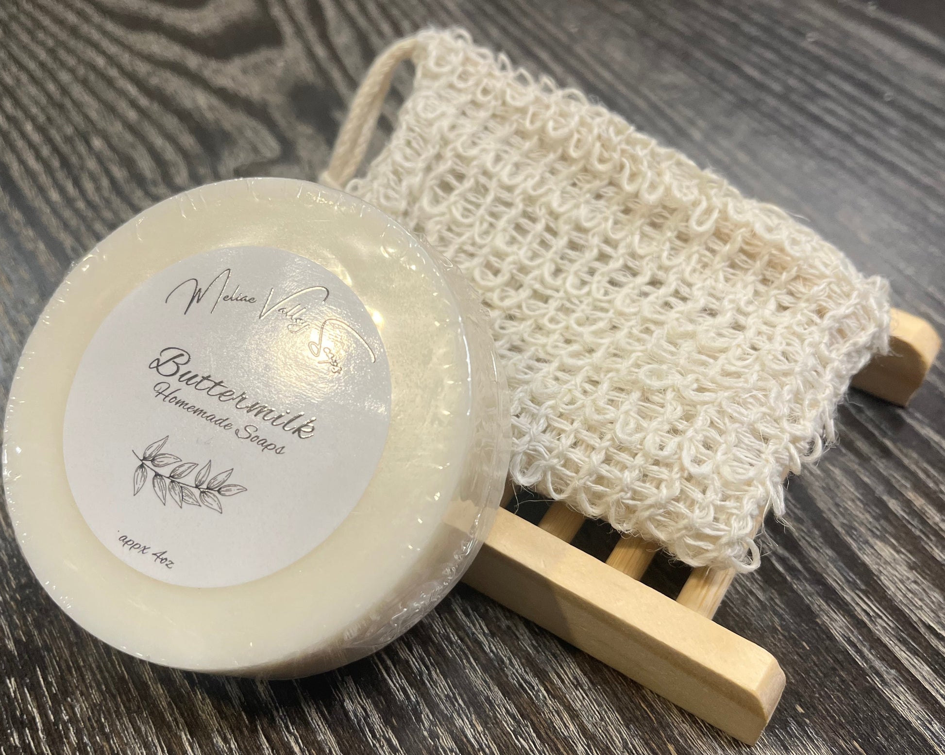 A round white soap with a wooden stand and an exfoliating bag on a dark wooden surface. 