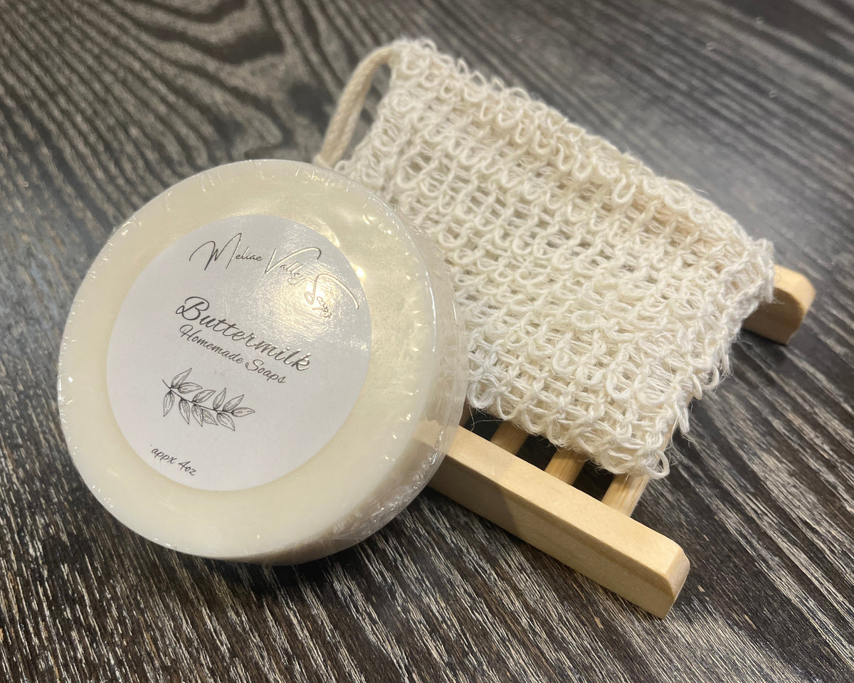 A white soap bar with a white sticker leans against an exfoliating bag and a soap bar holder. 