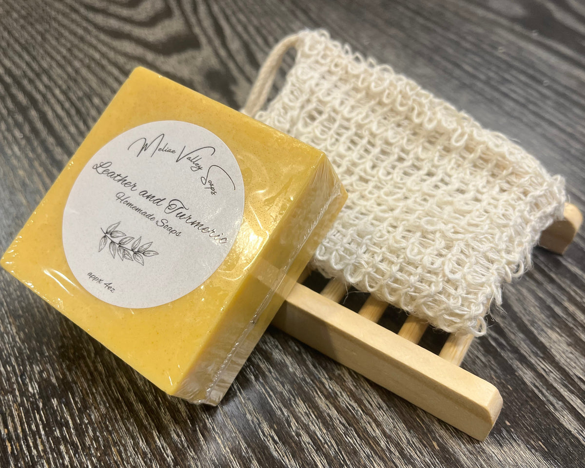 A yellowish-orange soap bar with a white sticker leans against an exfoliating bag and a soap bar holder. 