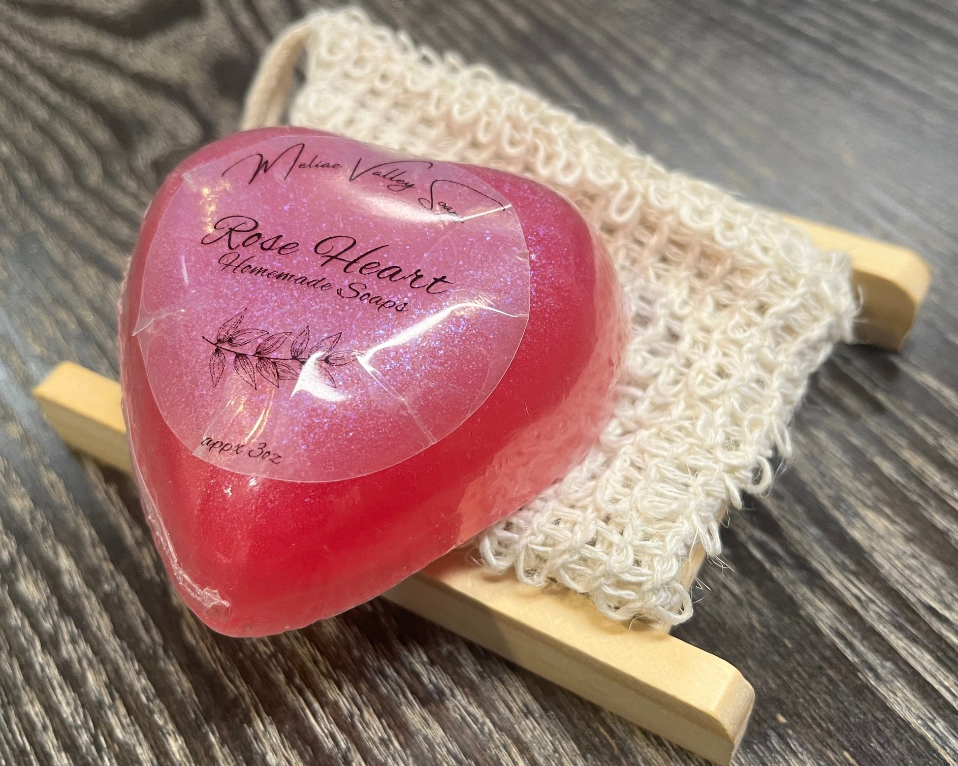 A pink heart-shaped bar of soap with a wooden stand and an exfoliating bag on a dark wooden surface. 
