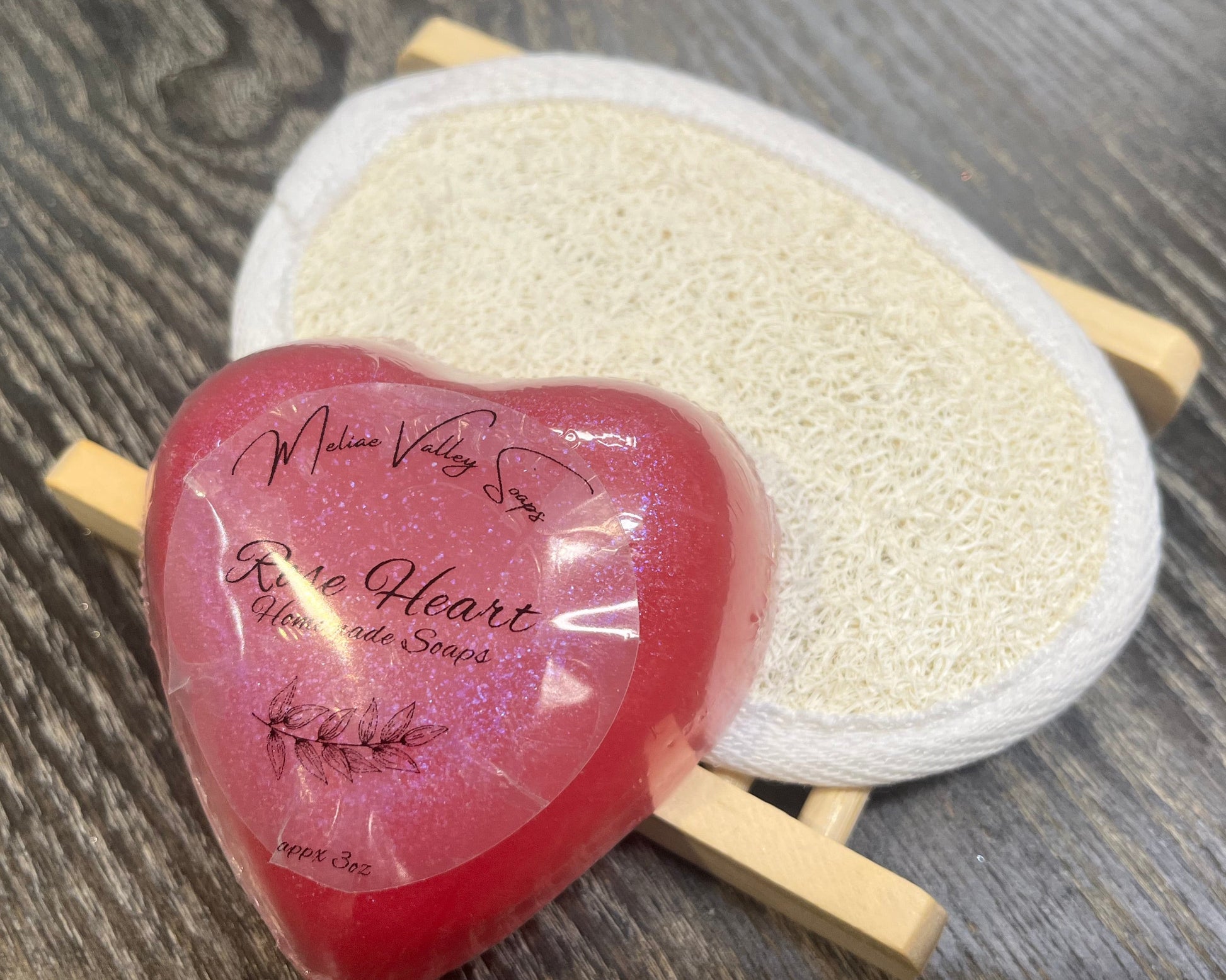 A pink heart-shaped bar of soap with a wooden stand and a loofah pad on a dark wooden surface. 