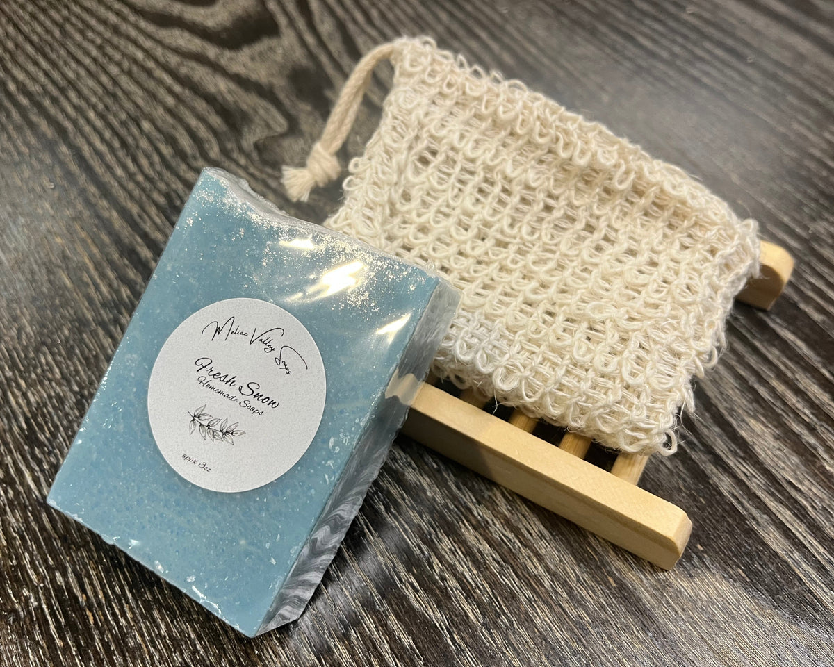 A blue soap bar with a white sticker leans against an exfoliating bag and a soap bar holder. 