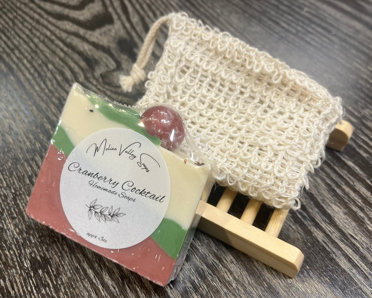 A white, red and green soap bar with a white sticker leans against an exfoliating bag and a soap bar holder. 