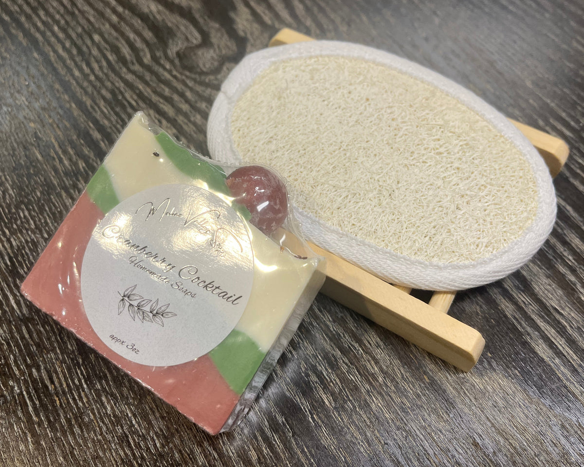 A white, red and green soap bar with a white sticker leans against a loofah pad and a soap bar holder.