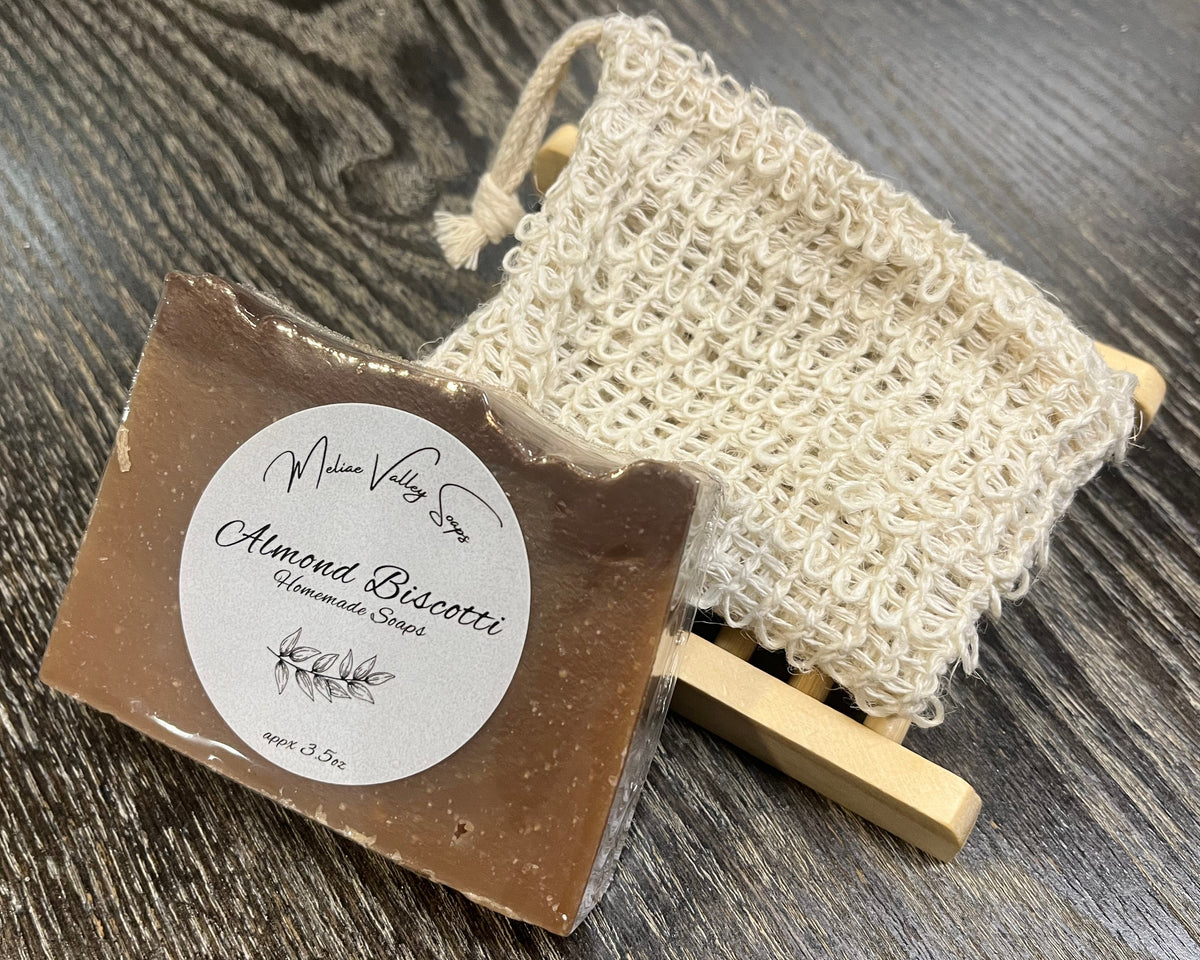 A brown soap bar with a white sticker leans against an exfoliating bag and a soap bar holder. 