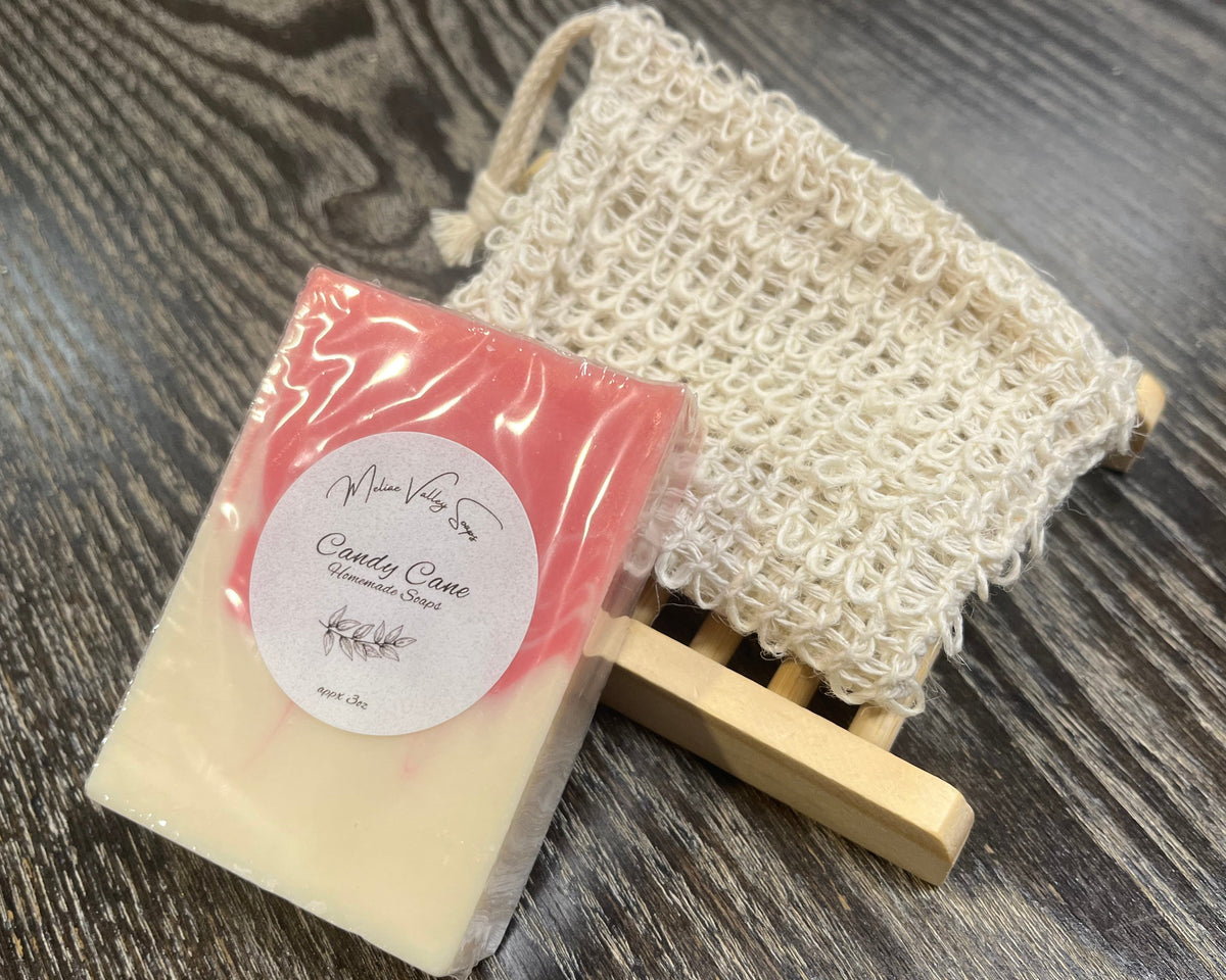 A beige and red soap bar with a white sticker leans against an exfoliating bag and a soap bar holder. 