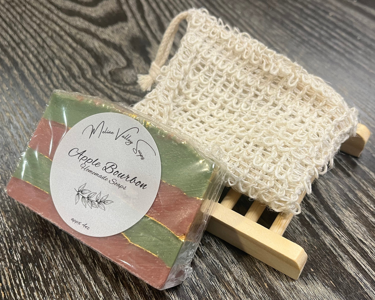 A red and green soap bar with a white sticker leans against an exfoliating bag and a soap bar holder. 