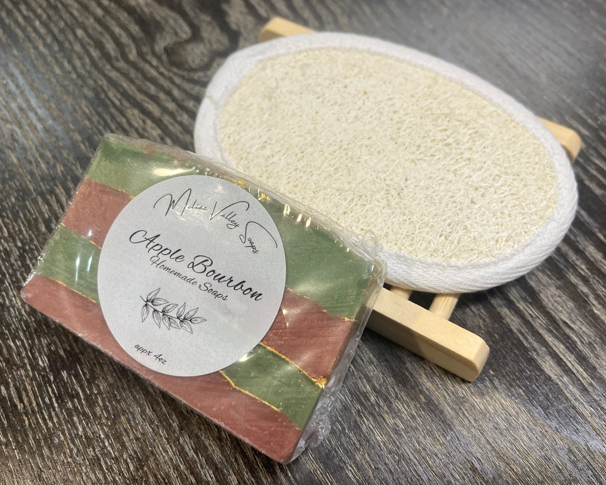 A red and green soap bar with a white sticker leans against a loofah pad and a soap bar holder. 