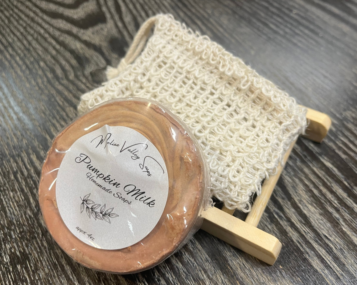 An orangey-brown soap bar with a white sticker leans against an exfoliating bag and a soap bar holder. 