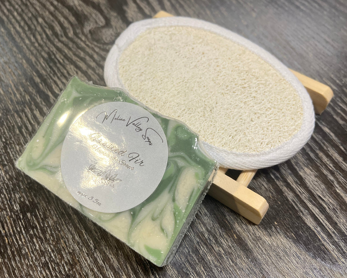 A white and green soap bar with a white sticker leans against a loofah pad and a soap bar holder.