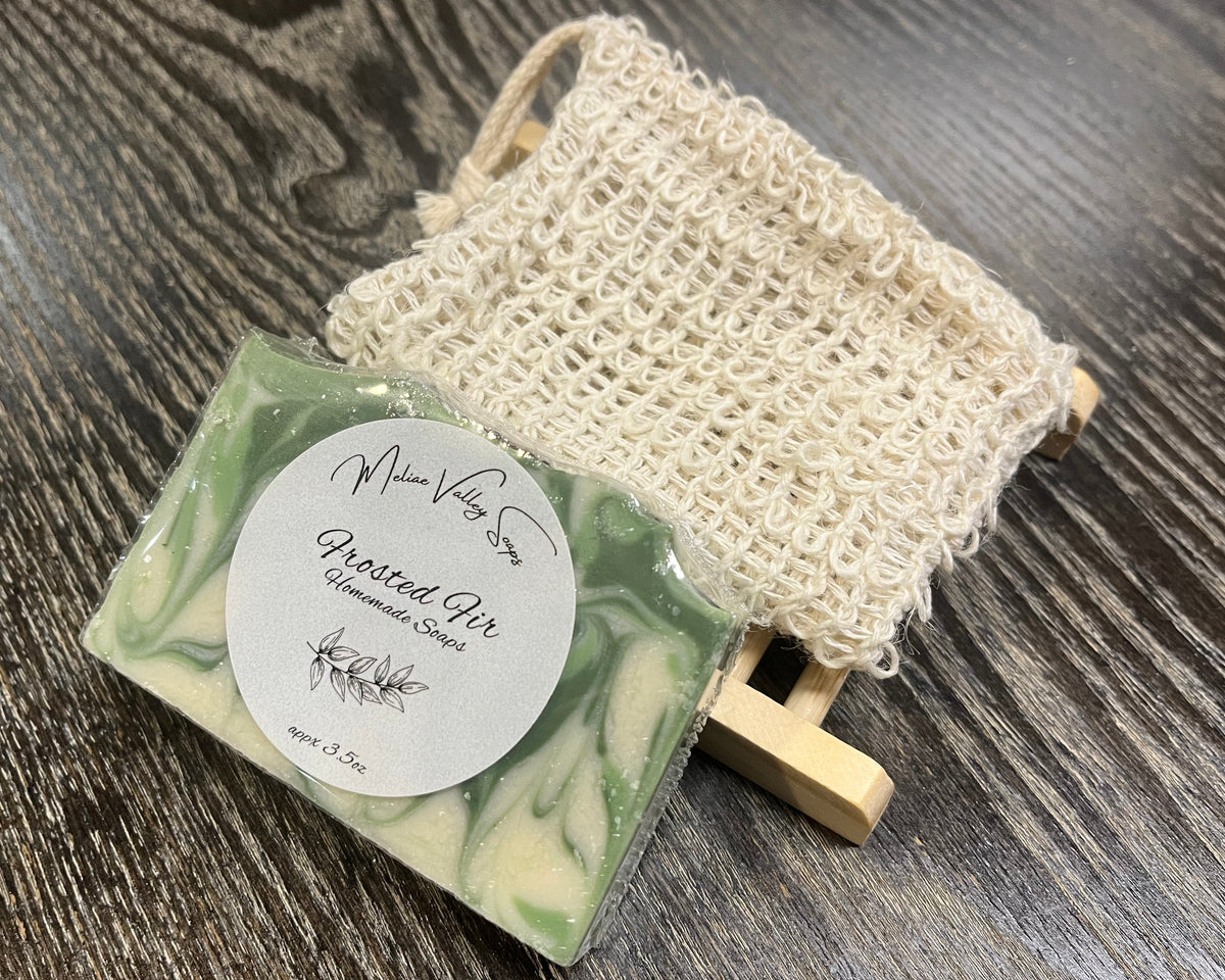A white and green soap bar with a white sticker leans against an exfoliating bag and a soap bar holder. 