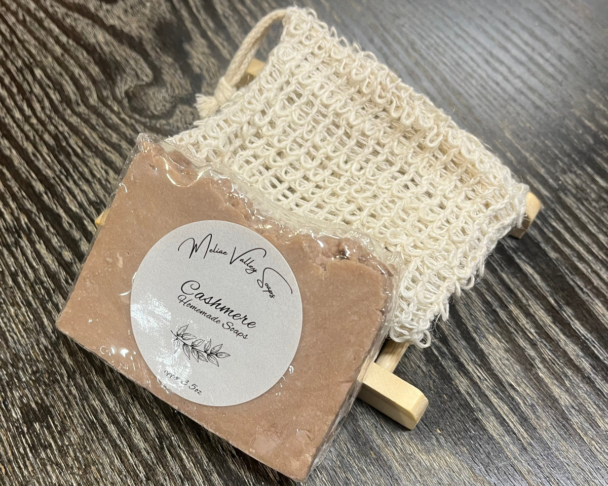 A pale purple soap bar with a white sticker leans against an exfoliating bag and a soap bar holder. 