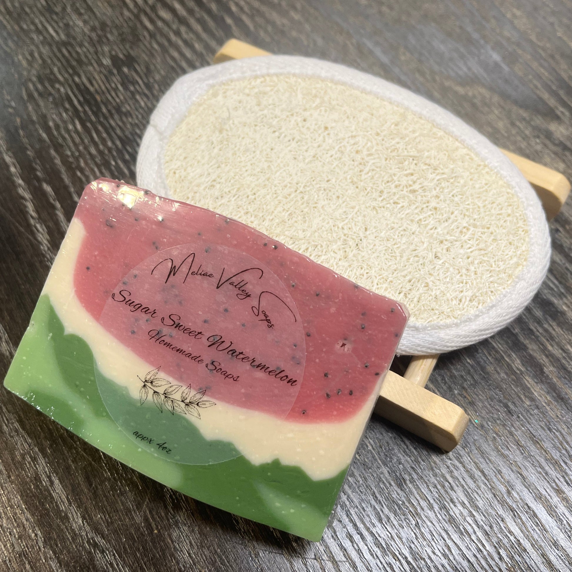 A watermelon-patterned soap with a wooden stand and a loofah pad on a dark wooden surface. 