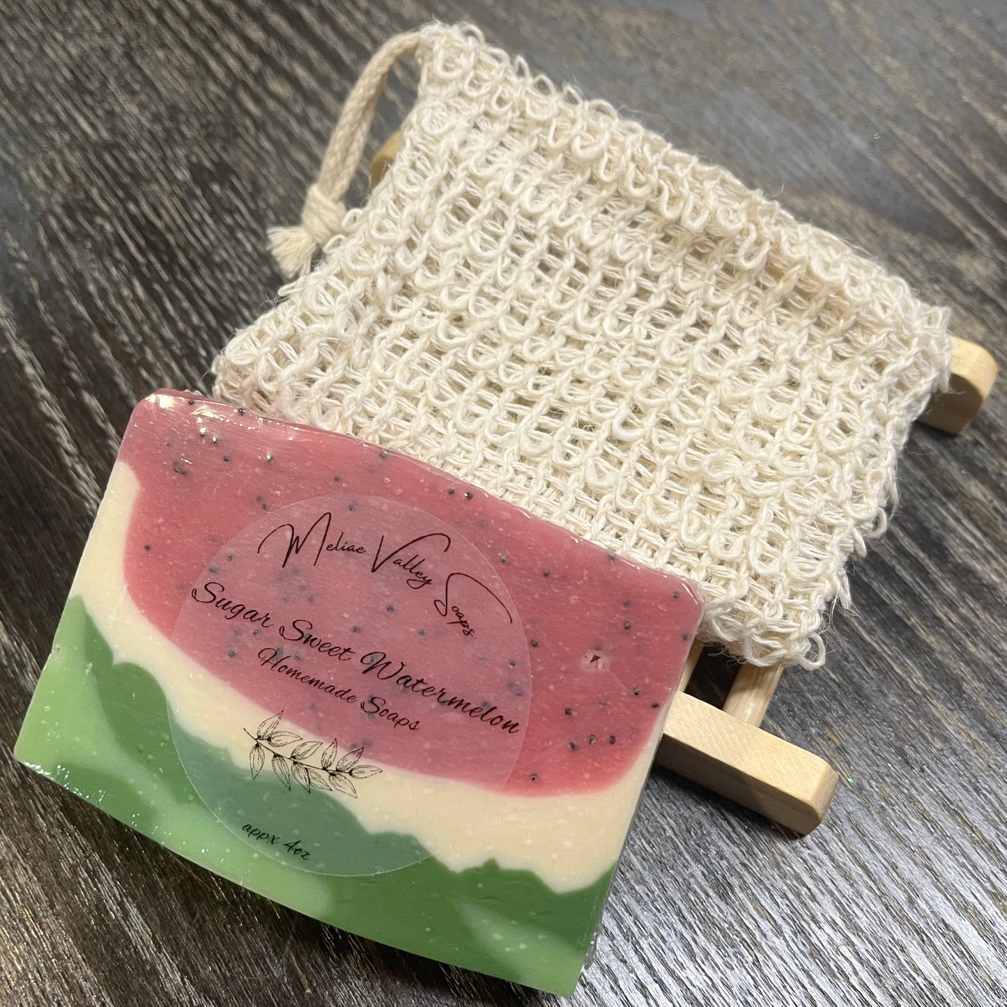 A watermelon-patterned soap with a wooden stand and an exfoliating bag on a dark wooden surface. 