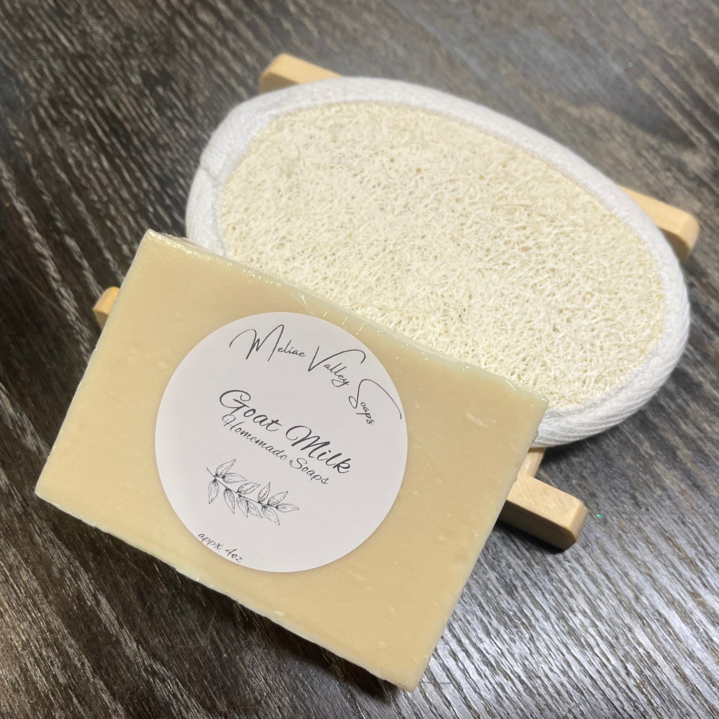A beige goat milk soap with a wooden stand and a loofah pad on a dark wooden surface. 
