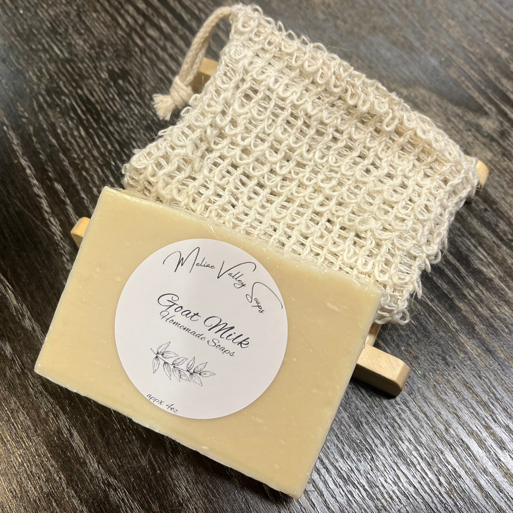 A beige goat milk soap with a wooden stand and an exfoliating bag on a dark wooden surface. 
