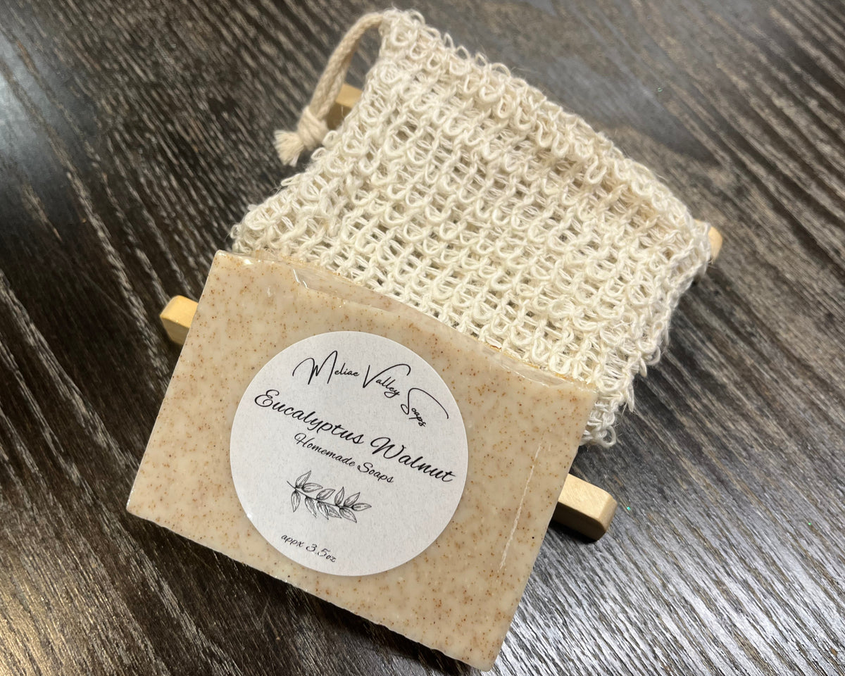 A beige soap bar with a white sticker leans against an exfoliating bag and a soap bar holder. 