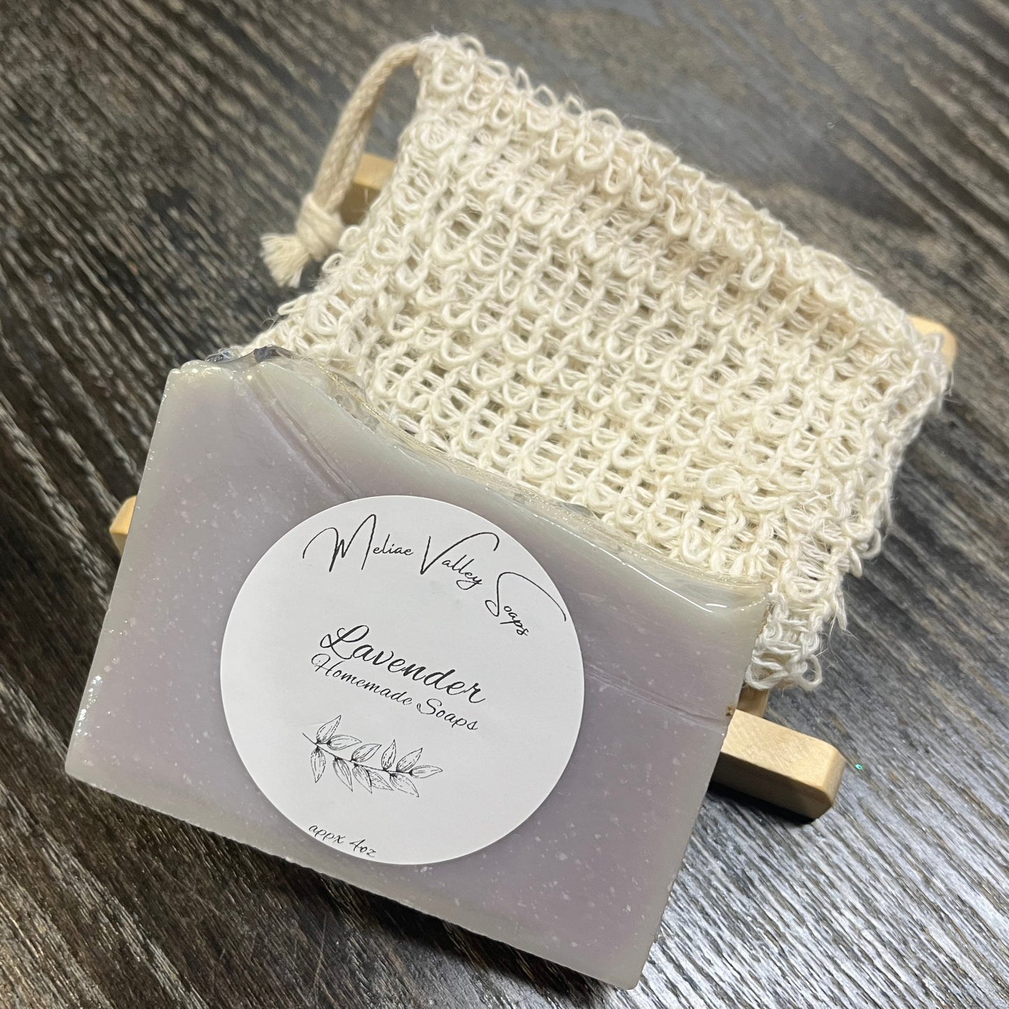 A purple bar of soap with a wooden stand and a exfoliating bag on a dark wooden surface. 