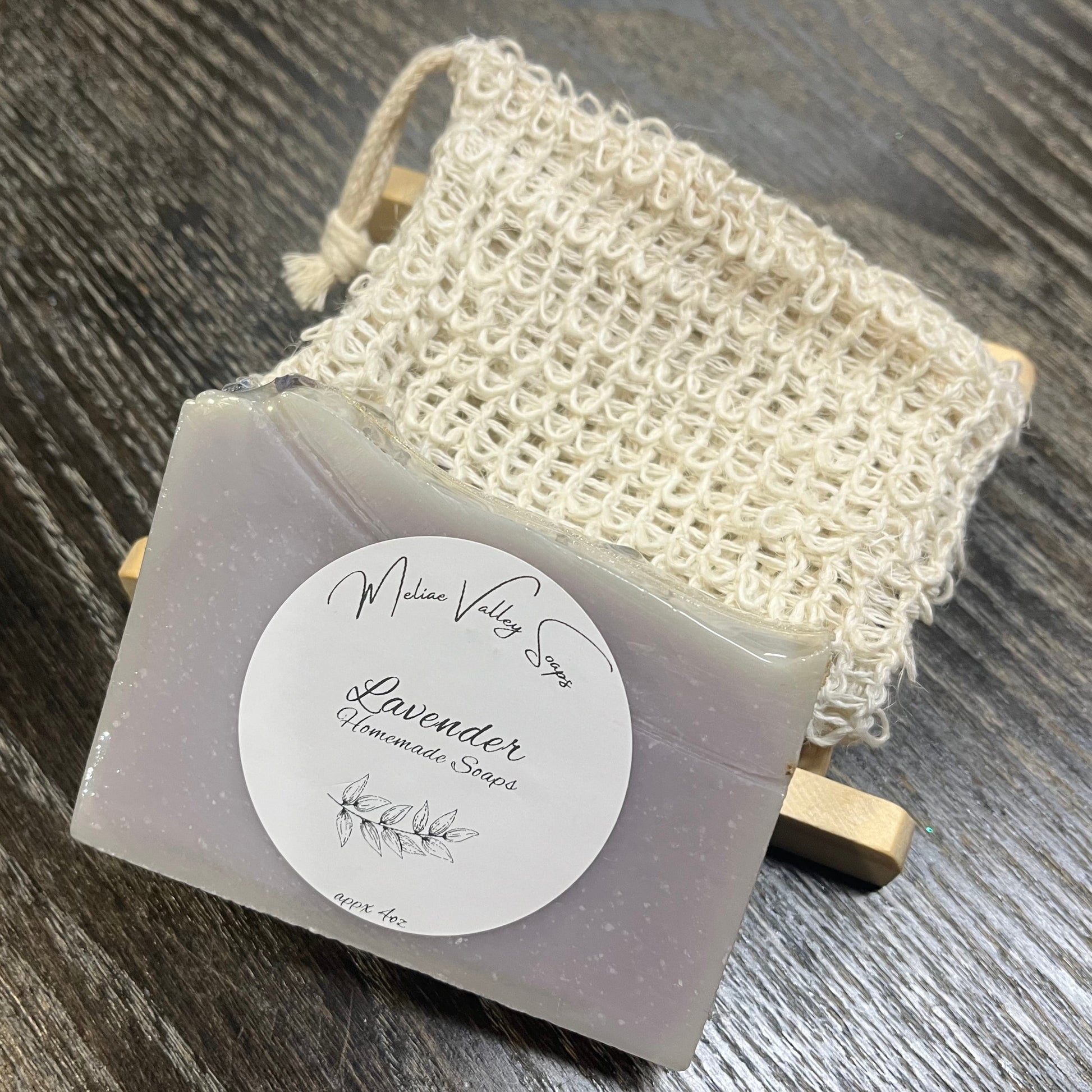 A purple bar of soap with a wooden stand and a exfoliating bag on a dark wooden surface. 