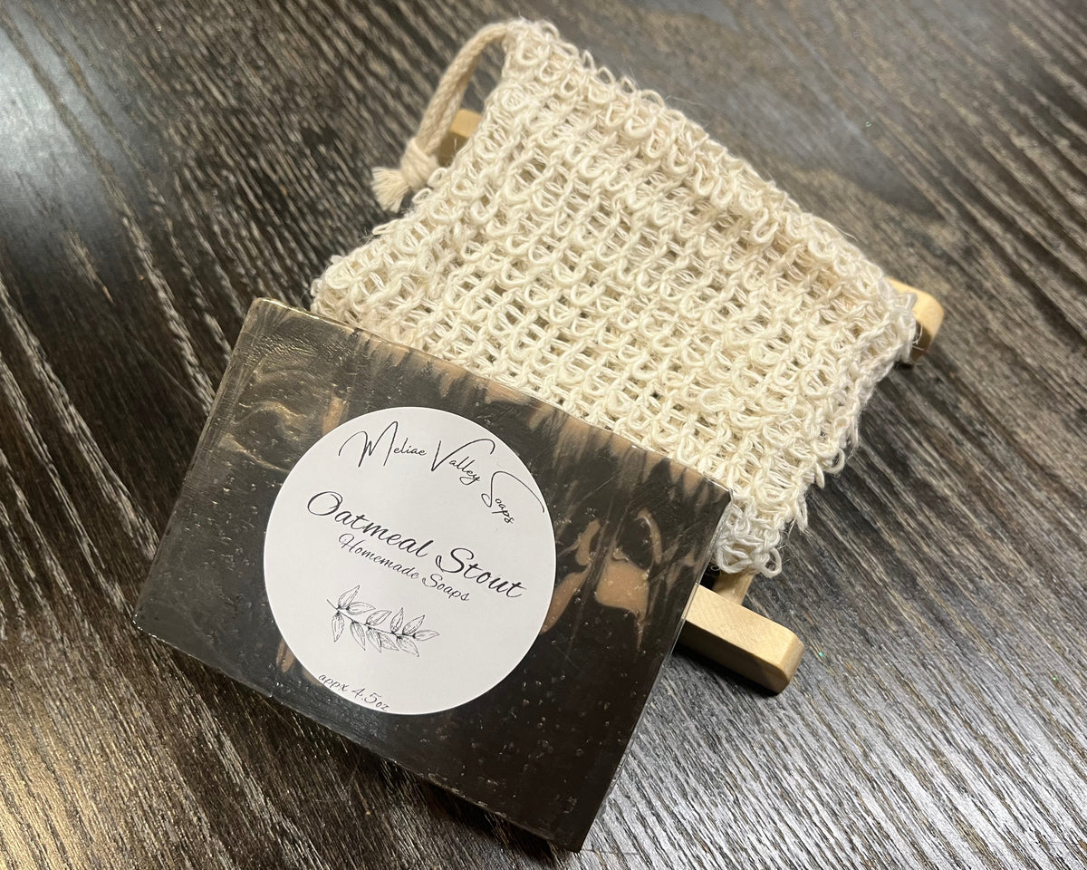 A beige and brown soap bar with a white sticker leans against an exfoliating bag and a soap bar holder. 