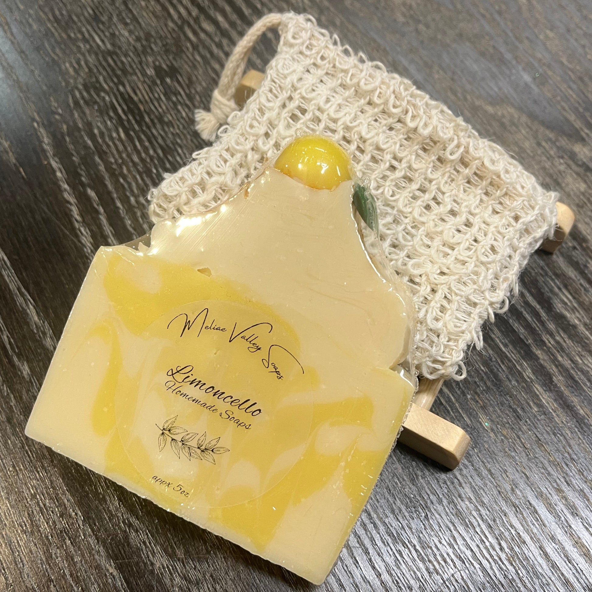 A yellow and white soap bar with a wooden stand and an exfoliating bag on a dark wooden surface. 