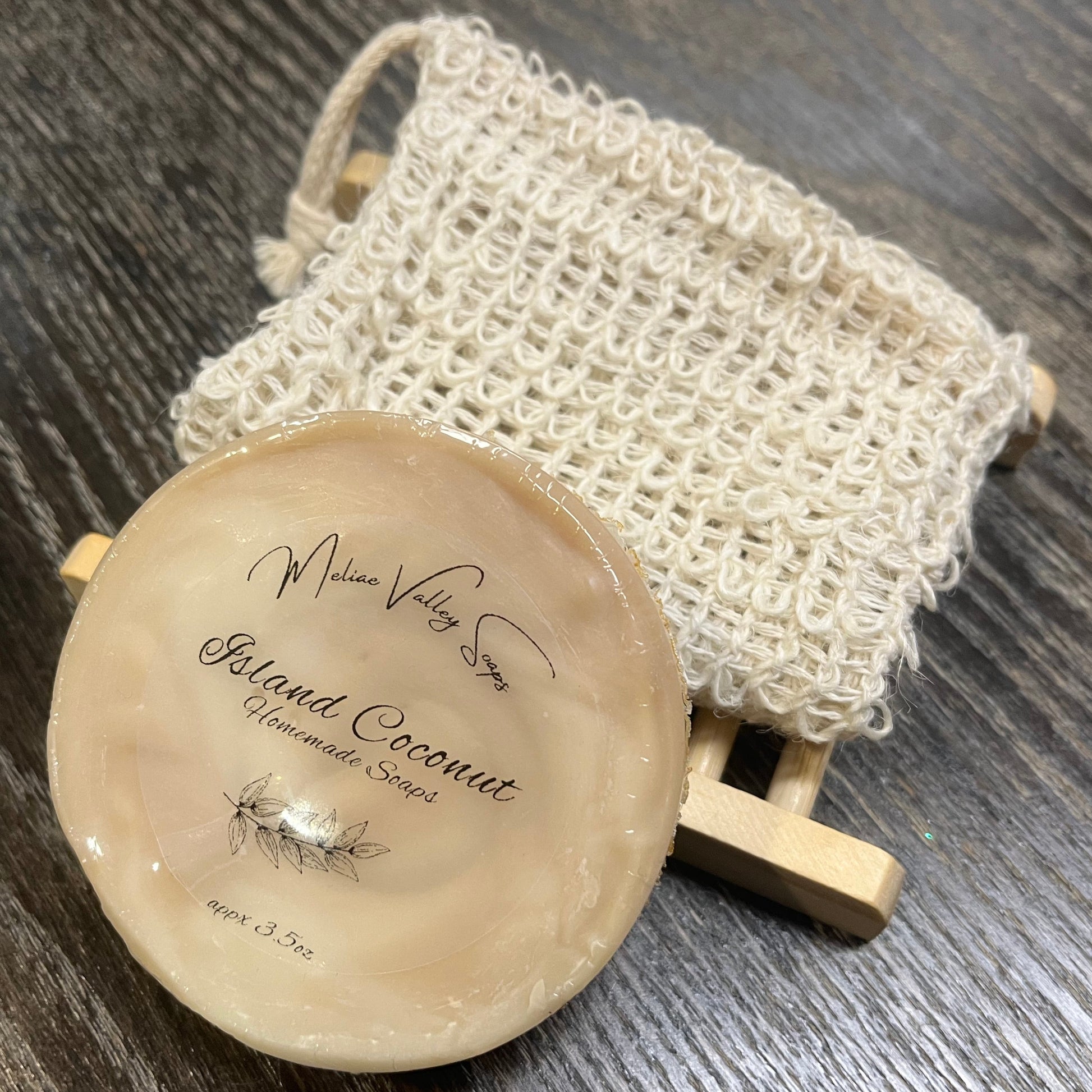 A round beige soap with a wooden stand and a exfoliating bag on a dark wooden surface. 