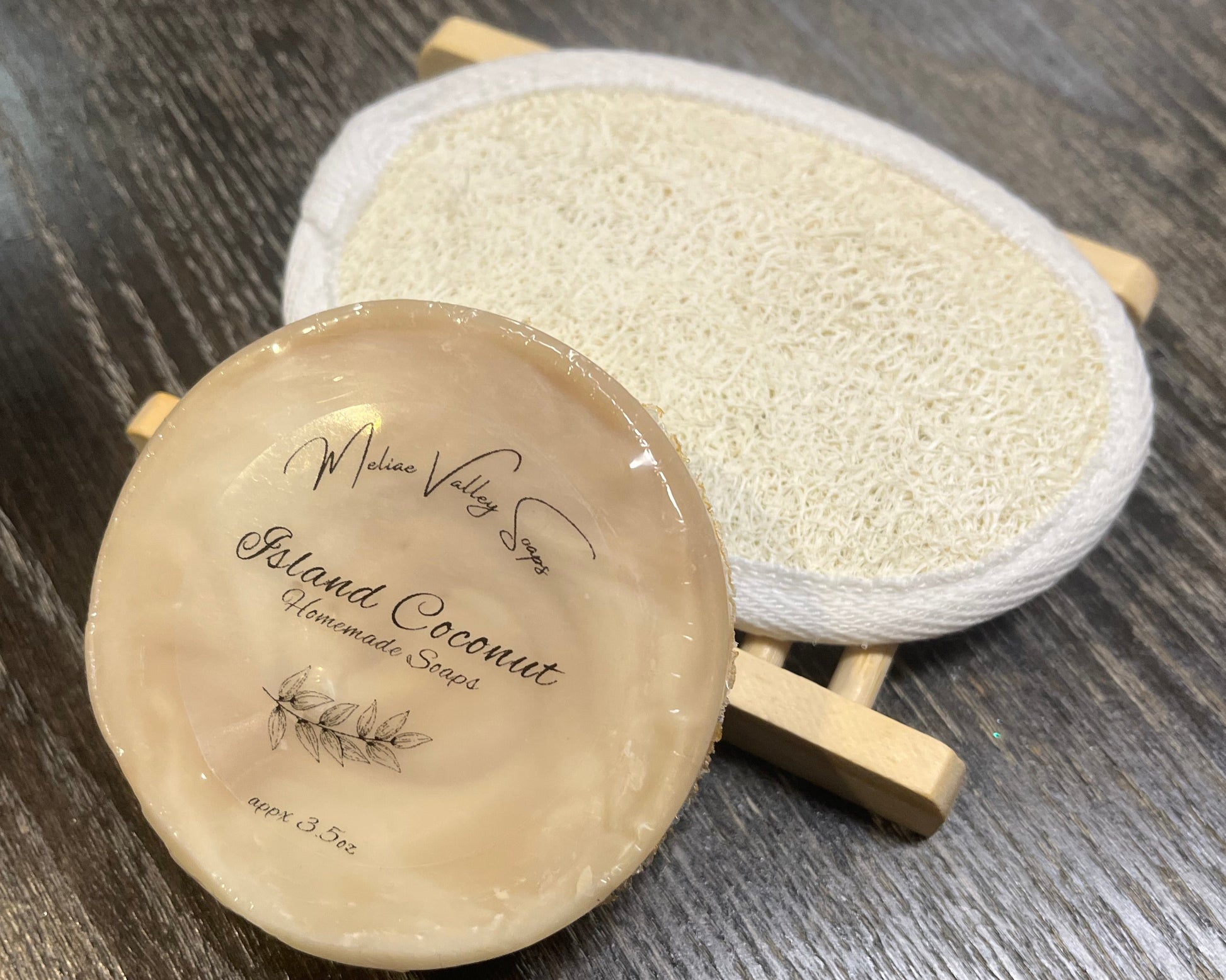 A round beige soap with a wooden stand and a loofah pad on a dark wooden surface. 