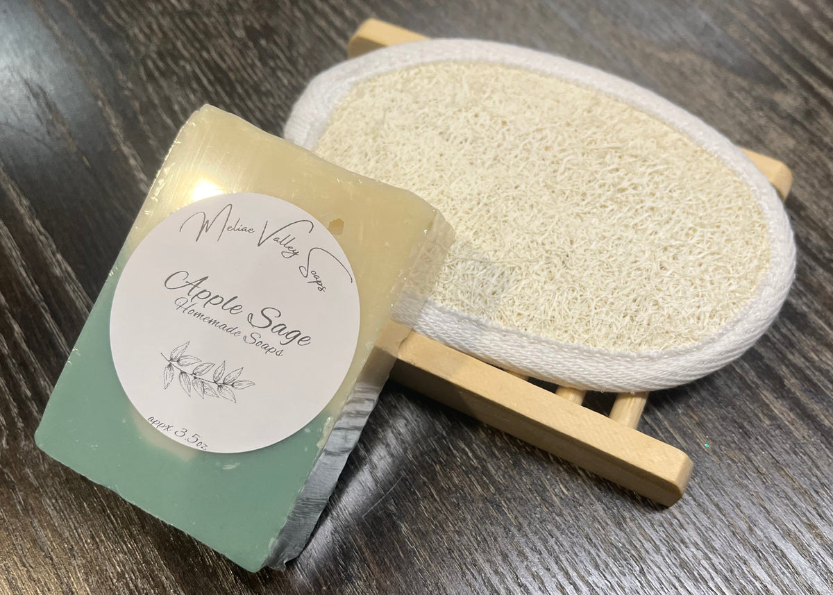 A beige and green soap bar with a white sticker leans against a loofah pad and a soap bar holder. 