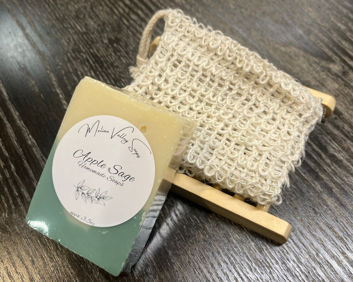 A beige and green soap bar with a white sticker leans against an exfoliating bag and a soap bar holder. 