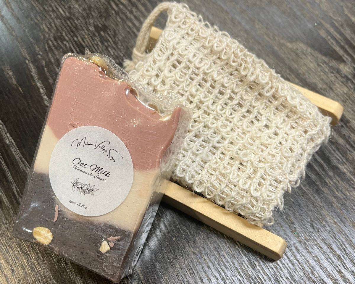 A beige, pink and brown soap bar with a white sticker leans against an exfoliating bag and a soap bar holder. 