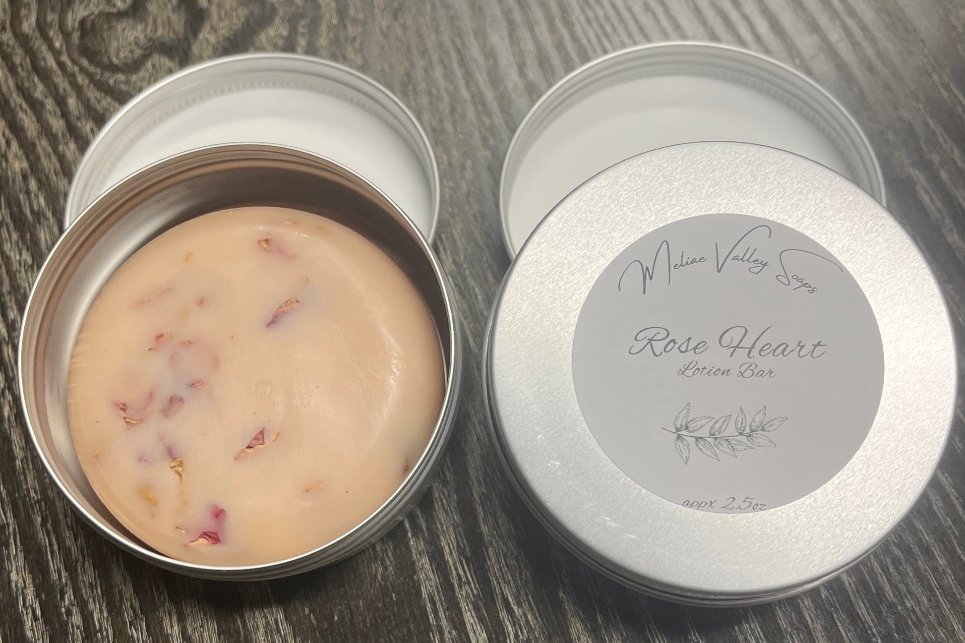 An open round metal tin showing a pink lotion bar with rose petals. Next to it is a silver lid with a white label on a dark wood-grain surface.
