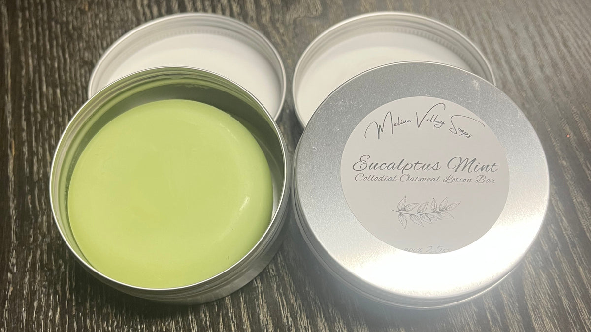 Two tins of lotion bars sit on a textured surface, with one showing a light green lotion bar in a metal tin and the other with a white label, both set against a blurred background.