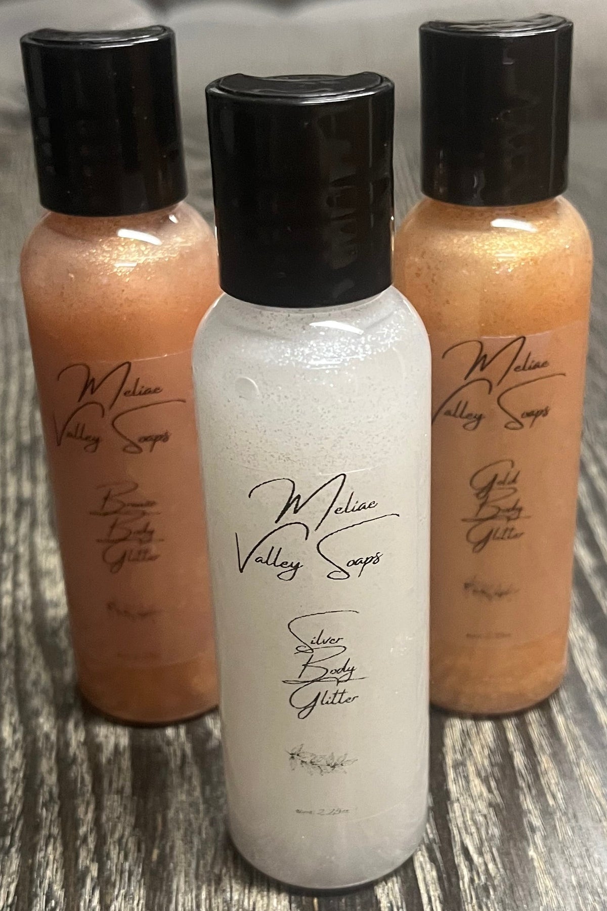 A triangle of three small bottles of body shimmer. The bottle in front is silver, the back-left bottle is bronze, and the back right bottle is gold. 