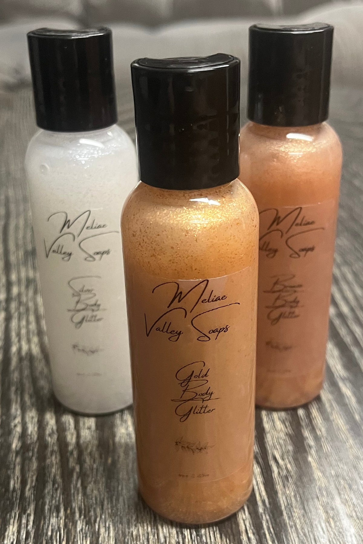 A triangle of three small bottles of body shimmer. The front bottle is gold, the back-left bottle is silver, and the back-right bottle is bronze. 