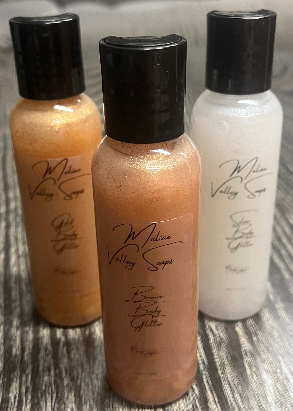 A triangle of three small bottles of body shimmer. The bottle in front is bronze, the back left bottle is gold, and the back right bottle is silver. 