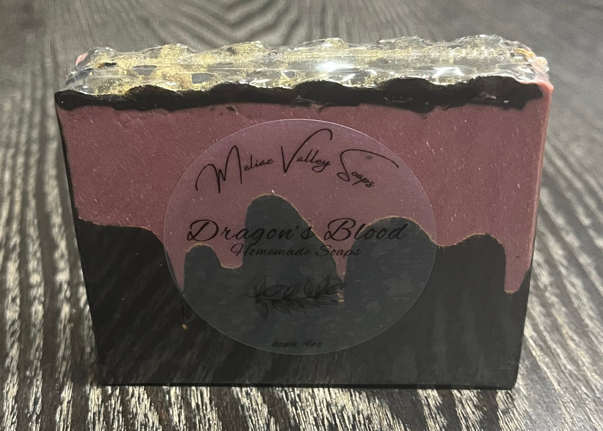 A bar of soap with a black bottom layer and a red layer made to look like blood. A thin black top layer is textured to look like gold dragon scales. There is a clear label on the front. 