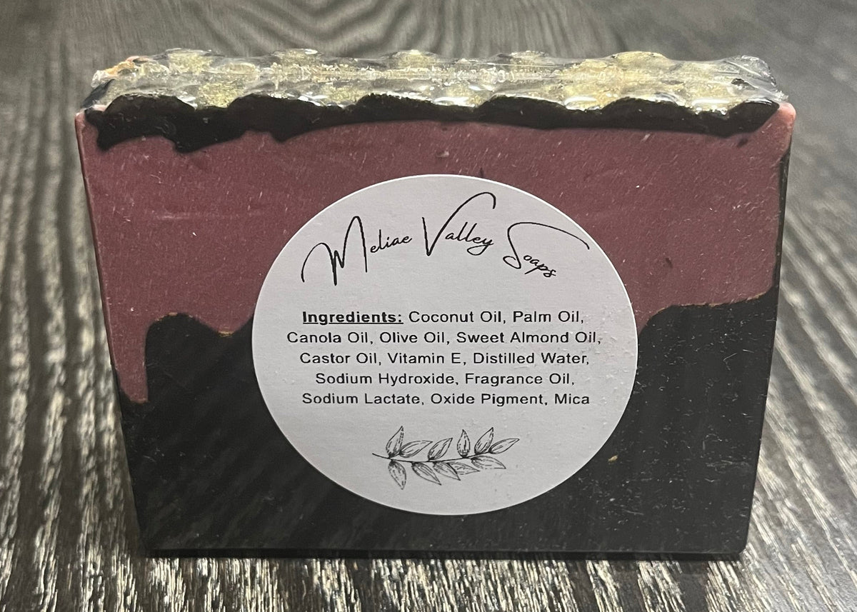 A bar of soap with a black bottom layer and a red layer made to look like blood. A thin black top layer is textured to look like gold dragon scales. There is a white ingredients label on it. 