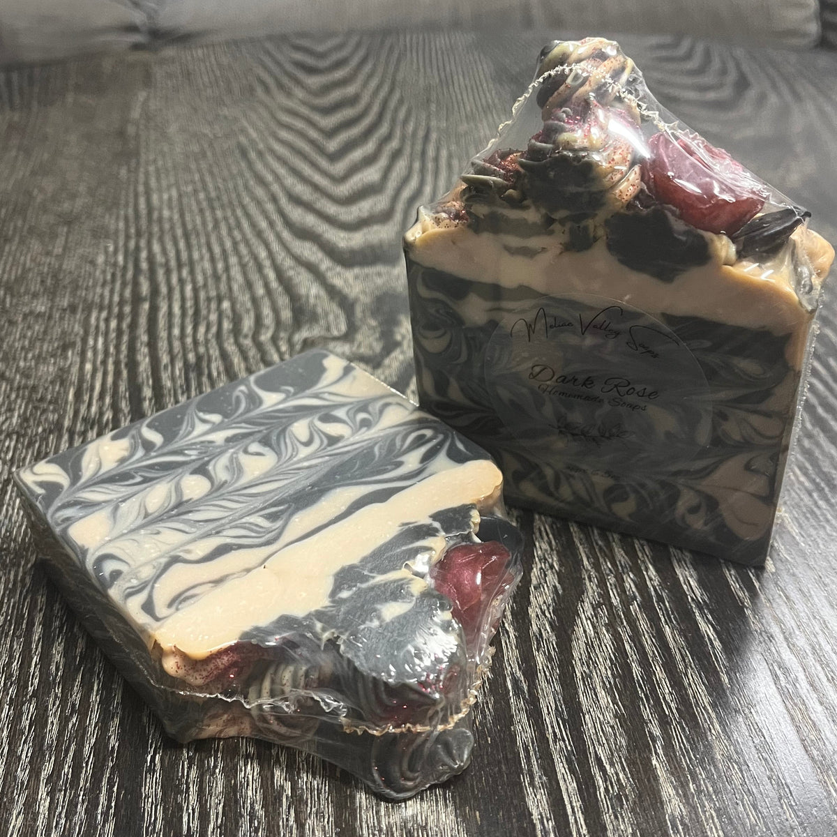 Two artisanal marbled soaps in clear plastic wrapping. The soaps are piled high with "frosting," with a dark red rose and a black leaf. 