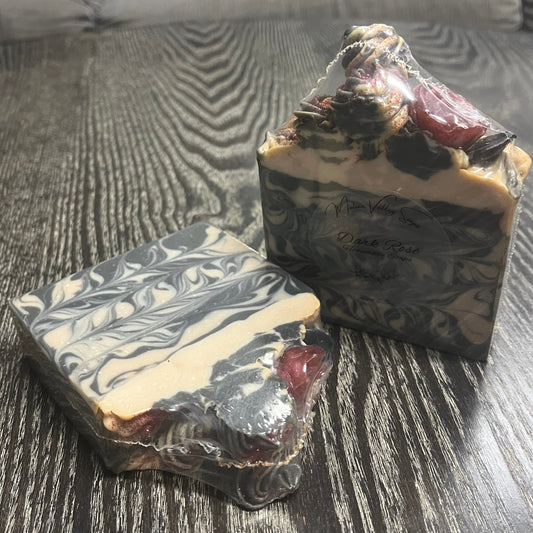 Two artisanal marbled soaps in clear plastic wrapping. The soaps are piled high with "frosting," with a dark red rose and a black leaf. 