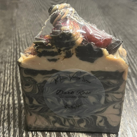 A rectangular bar of soap with a swirled black, gray, and cream pattern, topped with "soap frosting" and a small red rose. There is a clear label on the front. 