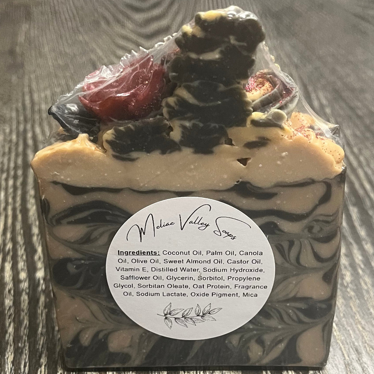A rectangular bar of soap with a swirled black, gray, and cream pattern, topped with "soap frosting" and a small red rose. There is a white ingredients label on it. 