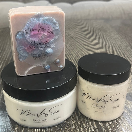 Two bottles next to each other. One has white whipped body butter; the other has an emulsified sugar scrub. On top of the body butter is a light gray bar of soap with a blue and purple orchid. All three have clear labels on the front. 