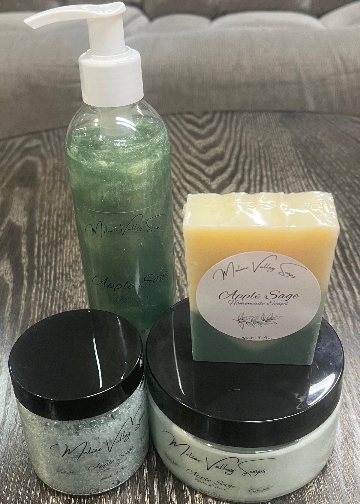 Two bottles next to each other. One has whipped body butter; the other has a sugar scrub. On top of the body butter is a bar of soap. Behind the sugar scrub is a bottle of liquid soap. All four have labels on the front. 