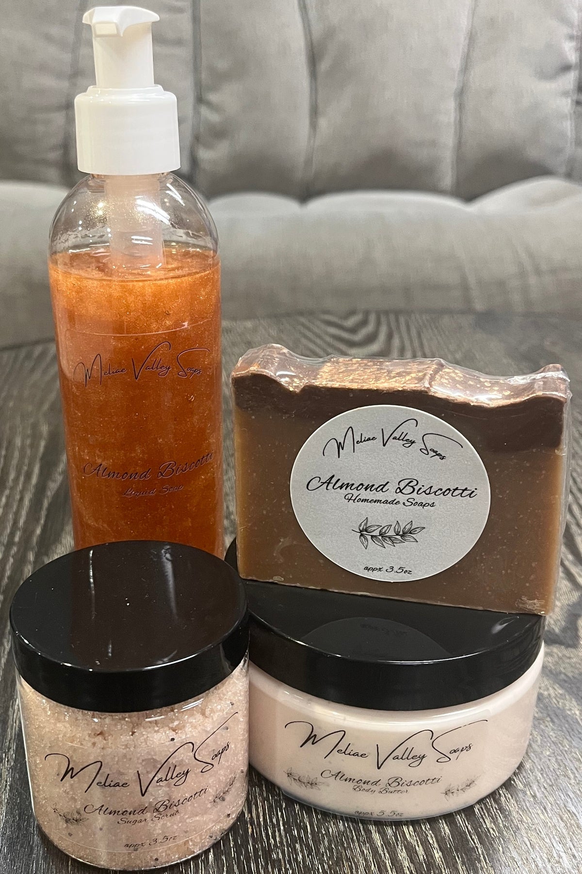 Two bottles next to each other. One has whipped body butter; the other has a sugar scrub. On top of the body butter is a bar of soap. Behind the sugar scrub is a bottle of liquid soap. All four have labels on the front. 