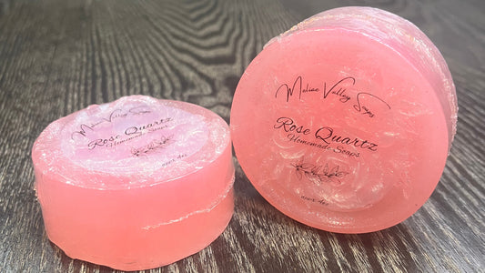 Two round bars of clear pink soap with designs on the front. The designs are dusted with white sparkles. The fronts of the bars have clear labels. 